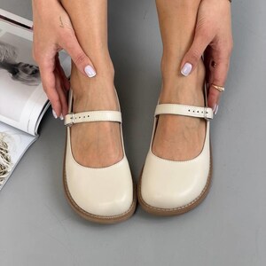 May include: Cream-coloured Mary Jane shoes with a strap and buckle. The shoes have a rounded toe and a brown sole. The shoes are made of a smooth material and are displayed on a grey surface.
