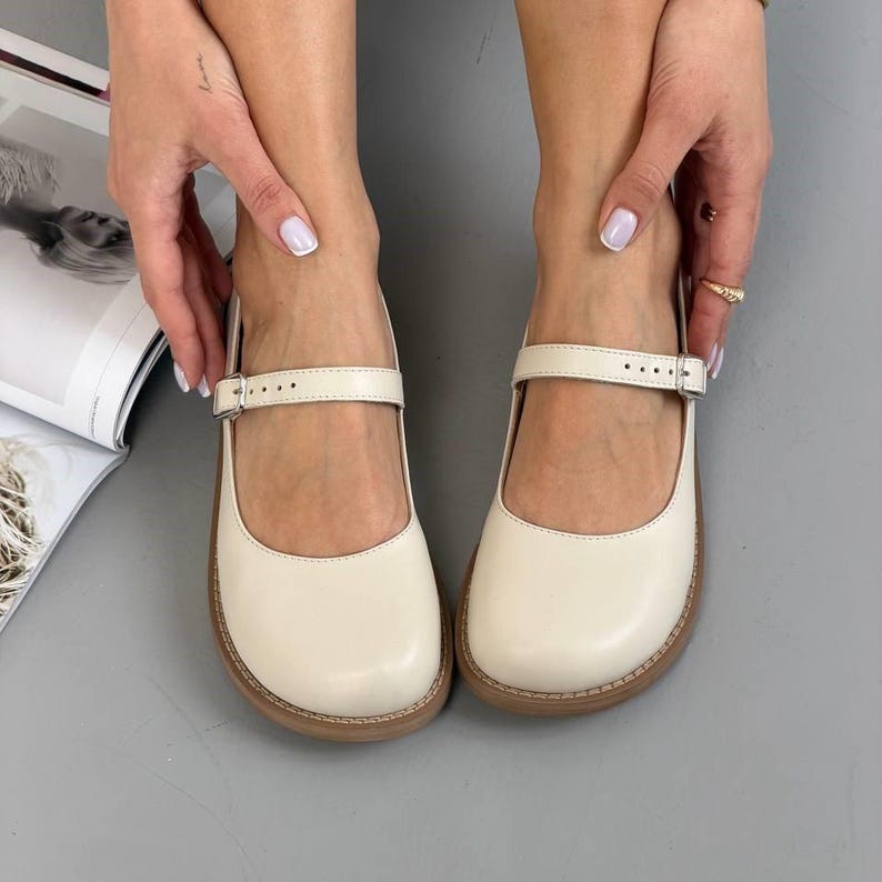 May include: Cream-coloured Mary Jane shoes with a strap and buckle. The shoes have a rounded toe and a brown sole. The shoes are made of a smooth material and are displayed on a grey surface.
