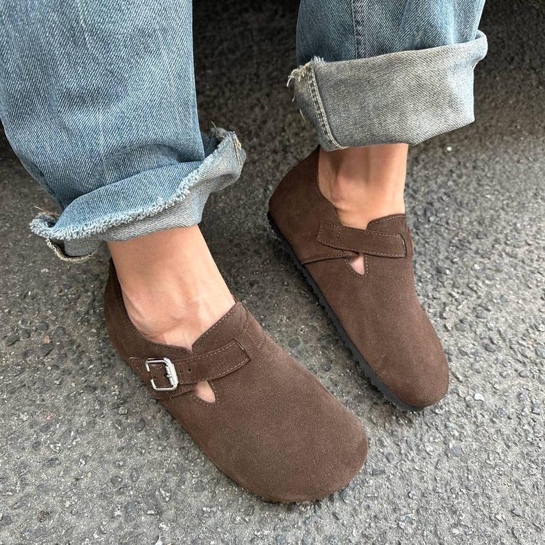 May include: Brown suede slip-on shoes with a buckle closure. The shoes have a rounded toe and a small cutout detail on the upper. The shoes are worn with blue jeans.