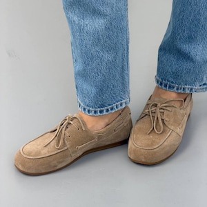 May include: Tan suede boat shoes with light brown laces. The shoes have a rounded toe and a stitched design around the edges. The shoes are worn with blue jeans.