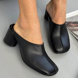 May include: Black leather mules with a square toe and block heel. The open-back design features a smooth, solid color, and a sturdy heel. These slip-on shoes are a stylish choice for everyday wear.