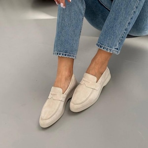May include: A pair of light beige suede loafers. The shoes have a classic slip-on design with a decorative strap across the top. The soles are a matching beige color. The shoes are shown on a gray surface.