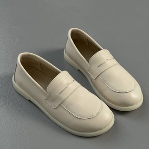May include: A pair of off-white leather loafers. The shoes have a rounded toe and a strap across the top. The soles are a matching off-white color. The shoes are displayed on a gray surface.