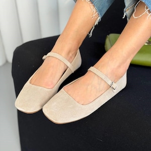 May include: Beige suede flats with a square toe and a delicate ankle strap. The shoes have a small buckle closure and a low heel. The flats are a neutral color, suitable for various outfits.