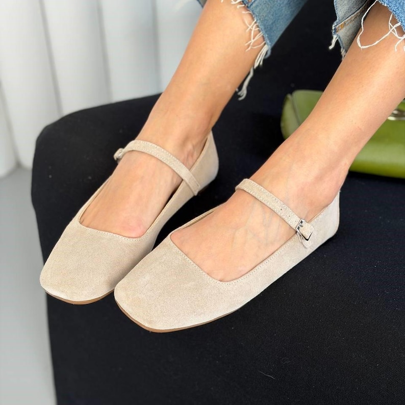 May include: Beige suede flats with a square toe and a delicate ankle strap. The shoes have a small buckle closure and a low heel. The flats are a neutral color, suitable for various outfits.