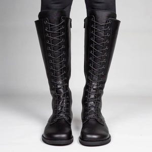May include: A pair of knee-high black boots featuring a full-length front lace-up design with dark eyelets and matching black laces. The boots have a rounded toe, a flat sole, and a side zip closure extending along the inner shaft of each boot.