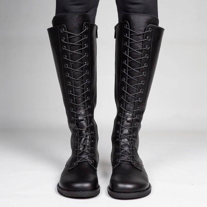 May include: A pair of knee-high black boots featuring a full-length front lace-up design with dark eyelets and matching black laces. The boots have a rounded toe, a flat sole, and a side zip closure extending along the inner shaft of each boot.