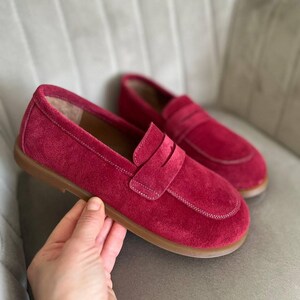 May include: A pair of red suede loafers with a penny strap. The shoes have a rounded toe and a light brown sole. The stitching is white, and the shoes are displayed on a neutral background.