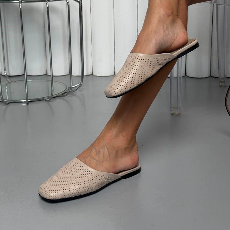May include: A pair of beige slip-on mules featuring a perforated texture across the upper surface. These shoes have a closed, rounded toe design and a flat sole, providing a simple and functional footwear option for casual or daily wear.