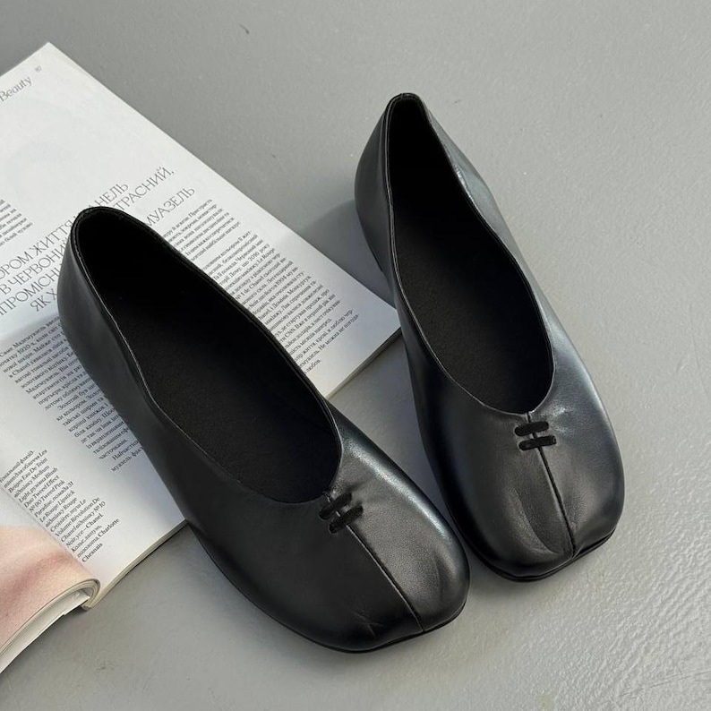 Op de afbeelding: Een paar zwarte leren ballerina's. De schoenen hebben een ronde neus en een klein decoratief detail op de wreef. De ballerina's worden gepresenteerd op een lichtgrijze ondergrond naast een open tijdschrift.