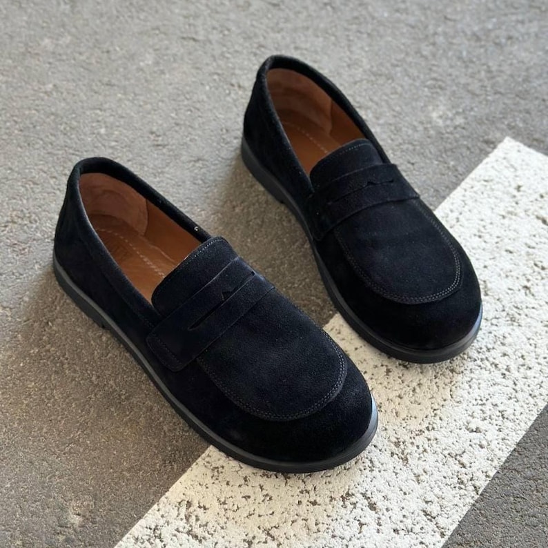 May include: A pair of black suede loafers. The shoes have a classic slip-on design with a rounded toe and a strap across the vamp. The soles are black, and the interior is a light brown color. The shoes are displayed on a gray and white surface.