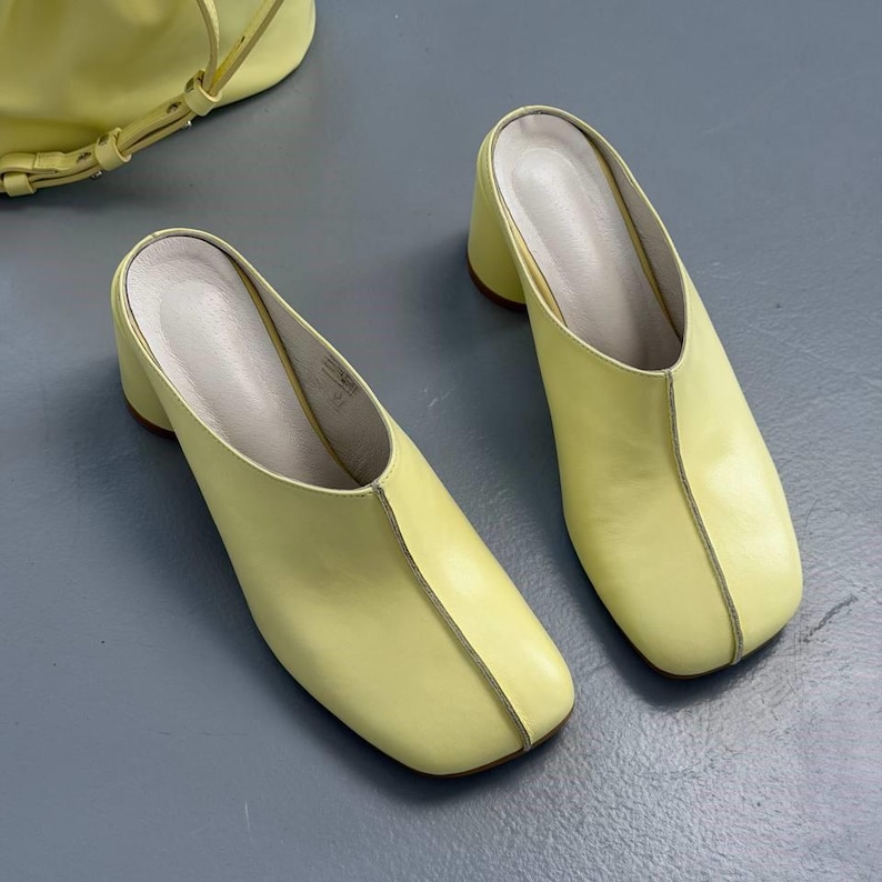 May include: A pair of pale yellow leather mules with a square toe and a block heel. The shoes have a minimalist design with a stitched seam down the center. A matching yellow handbag is visible in the background.