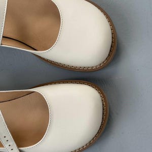 May include: Close-up of two white Mary Jane shoes. The shoes have a rounded toe, a strap, and a brown sole with visible stitching. The interior of the shoe is a light brown colour.