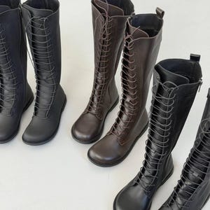 May include: A collection of knee-high boots featuring a lace-up front design with matching laces, rounded toes, and side zippers. The boots are available in solid black and dark brown colors, constructed with a smooth finish.