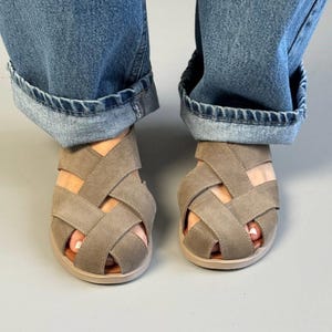May include: A pair of taupe-colored sandals with a woven strap design. The sandals have a rounded toe and a light brown sole. The image shows the sandals worn with blue denim jeans.