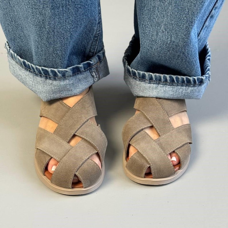 May include: A pair of taupe-colored sandals with a woven strap design. The sandals have a rounded toe and a light brown sole. The image shows the sandals worn with blue denim jeans.