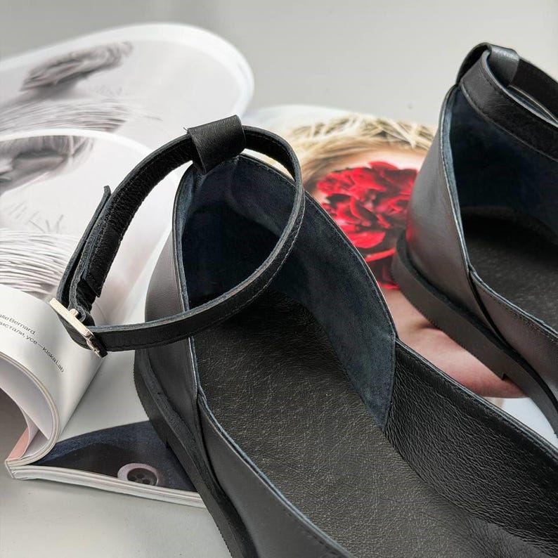 May include: Close-up of a pair of black leather flats with ankle straps. The shoes have a simple design with a rounded toe and a small heel. The ankle straps fasten with a silver-colored buckle. The shoes are on top of a magazine.