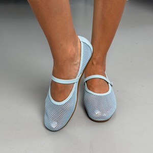 May include: A pair of light blue Mary Jane flats featuring a mesh upper construction with a fine grid pattern. The shoes include a thin, matching light blue strap across the instep with a small buckle closure and a rounded toe design.