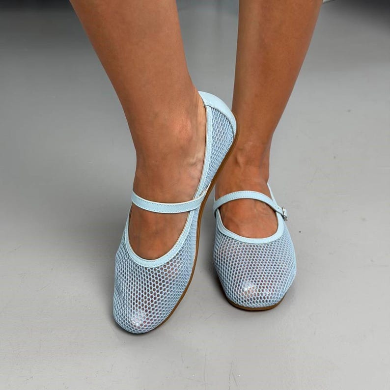May include: A pair of light blue Mary Jane flats featuring a mesh upper construction with a fine grid pattern. The shoes include a thin, matching light blue strap across the instep with a small buckle closure and a rounded toe design.