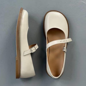 May include: Two off-white Mary Jane shoes with a square toe and a strap that buckles across the top. They have a light brown sole and a low heel. The shoes are made of a smooth, leather-like material. The shoes are approximately 23 cm long.
