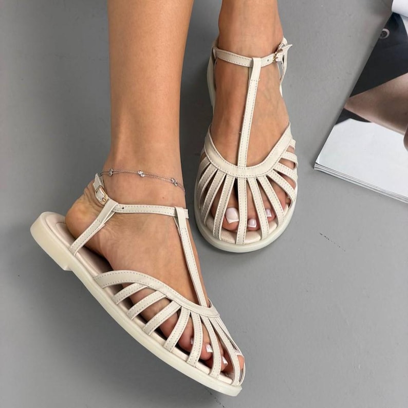 Puede incluir: Sandalias de cuero color crema con dise&ntilde;o de tira en T y m&uacute;ltiples correas en el pie. Las sandalias tienen suela plana y correas ajustables en el tobillo. El calzado se muestra sobre una superficie gris.