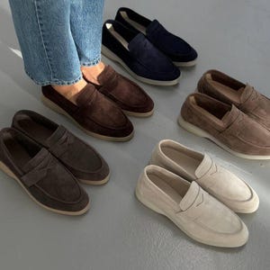 May include: A collection of suede penny loafers in various colors, including navy blue, brown, and beige. The shoes have a classic design with a rounded toe and a strap across the vamp. The loafers are displayed on a gray surface.