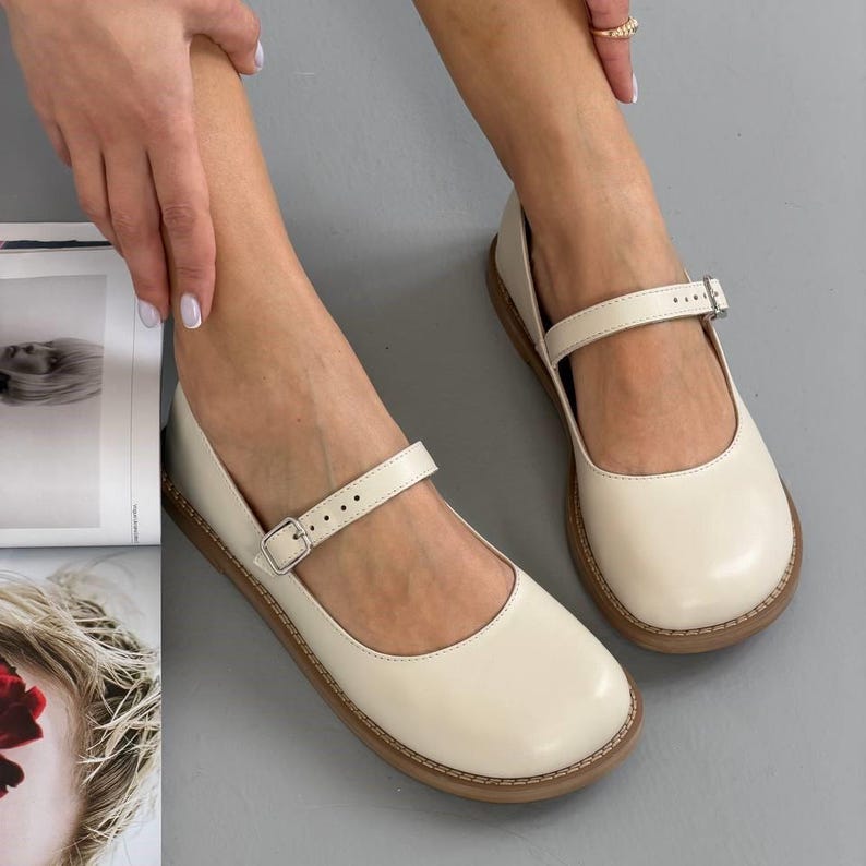 May include: Cream-coloured Mary Jane style shoes with a strap and buckle closure. The shoes have a rounded toe and a brown sole. The shoes are made of a smooth material and are shown on a grey surface.