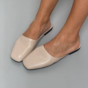 May include: A pair of beige slip-on mules featuring a square toe design and a perforated upper surface. The footwear has a low-profile construction with a thin black sole, providing a clean and simple aesthetic for everyday wear and casual use.