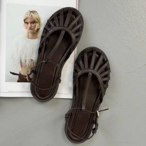 Puede incluir: Un par de sandalias de cuero marr&oacute;n oscuro con un dise&ntilde;o similar a una jaula. Las sandalias tienen una correa en T y una correa en el tobillo con una hebilla dorada. Las sandalias son planas y parecen nuevas.