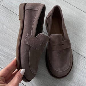May include: A pair of taupe suede loafers featuring a classic penny slot strap across the vamp, a rounded toe, and a flat brown sole. The shoes have a soft, matte texture and a simple slip-on design with visible stitching along the edges.