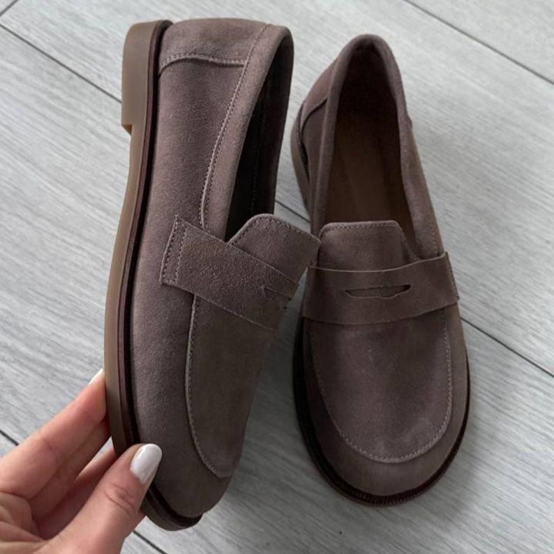 May include: A pair of taupe suede loafers featuring a classic penny slot strap across the vamp, a rounded toe, and a flat brown sole. The shoes have a soft, matte texture and a simple slip-on design with visible stitching along the edges.