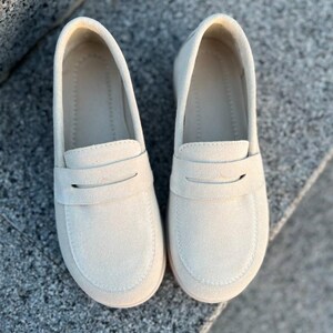 May include: A pair of cream-colored loafers. These slip-on shoes feature a rounded toe, a strap across the vamp, and a low heel. The shoes are made of a soft, suede-like material and are designed for comfort and style.