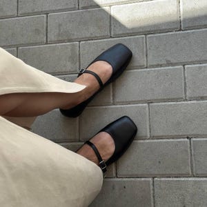May include: Black Mary Jane style flats with a square toe and a strap that buckles across the top of the foot. The shoes are on a gray brick surface. The person wearing the shoes is wearing beige pants.