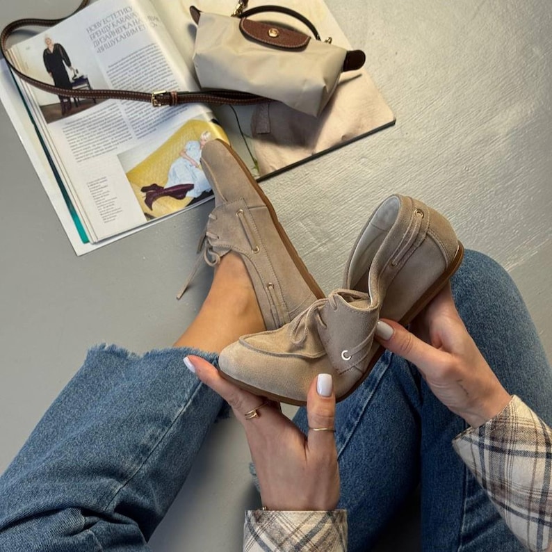 May include: A pair of beige suede boat shoes with laces, displayed with a matching handbag and a magazine. The shoes are a light tan color and have a classic design. The handbag is a light beige with a brown leather flap.