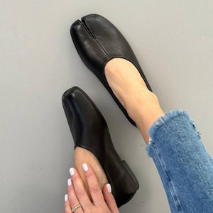 May include: Black leather split-toe flats. The shoes have a square toe design and a low heel. The design features a split between the big toe and the other toes. The shoes are shown from an overhead perspective.