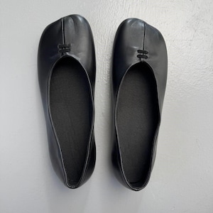 Op de afbeelding: Een paar zwarte leren ballerina's. De schoenen hebben een split-teenontwerp en een klein decoratief detail aan de bovenkant. De zolen zijn donker en de schoenen lijken ongedragen.