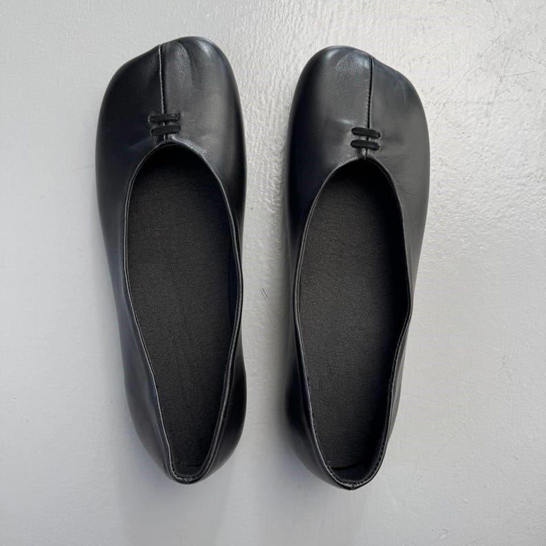 Op de afbeelding: Een paar zwarte leren ballerina's. De schoenen hebben een split-teenontwerp en een klein decoratief detail aan de bovenkant. De zolen zijn donker en de schoenen lijken ongedragen.