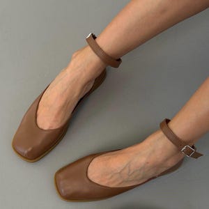 May include: Brown leather flats with a square toe and ankle strap. The flats have a buckle closure and a simple, elegant design. The shoes are a warm brown color and appear to be made of smooth leather.