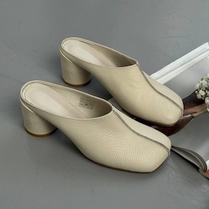 Puede incluir: Mules de cuero color crema con punta cuadrada y tacón ancho. Los zapatos tienen una superficie texturizada y una costura central. Los tacones miden unos 7.6 cm de alto. Calzado de moda.