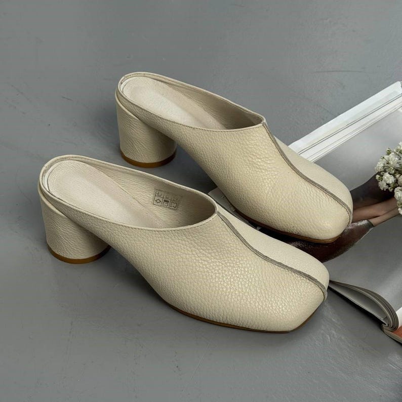 May include: A pair of cream-colored leather mules with a square toe and block heel. The shoes have a slip-on design and a textured surface. The heels are approximately 3 inches high.