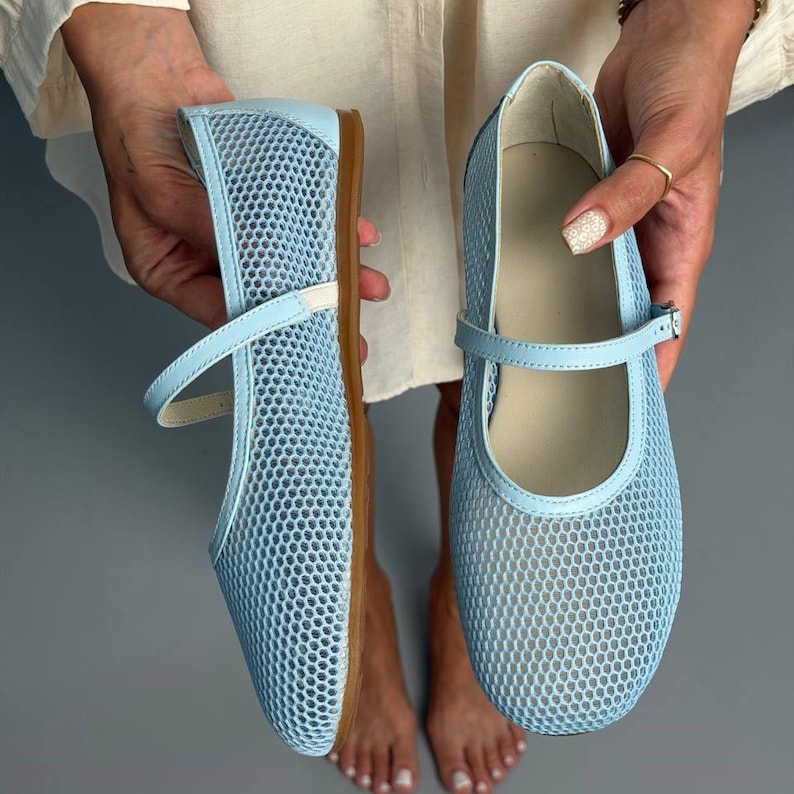 May include: A pair of light blue Mary Jane flats featuring a breathable mesh upper, a rounded toe, and a thin adjustable strap with a buckle closure. The shoes have a flat sole and a light-coloured interior lining, showcasing a simple design.