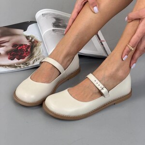 May include: Cream Mary Jane shoes with a rounded toe and a strap with a buckle. The shoes have a flat sole with a light brown trim. The shoes are displayed on a grey surface. The shoes are a classic style.