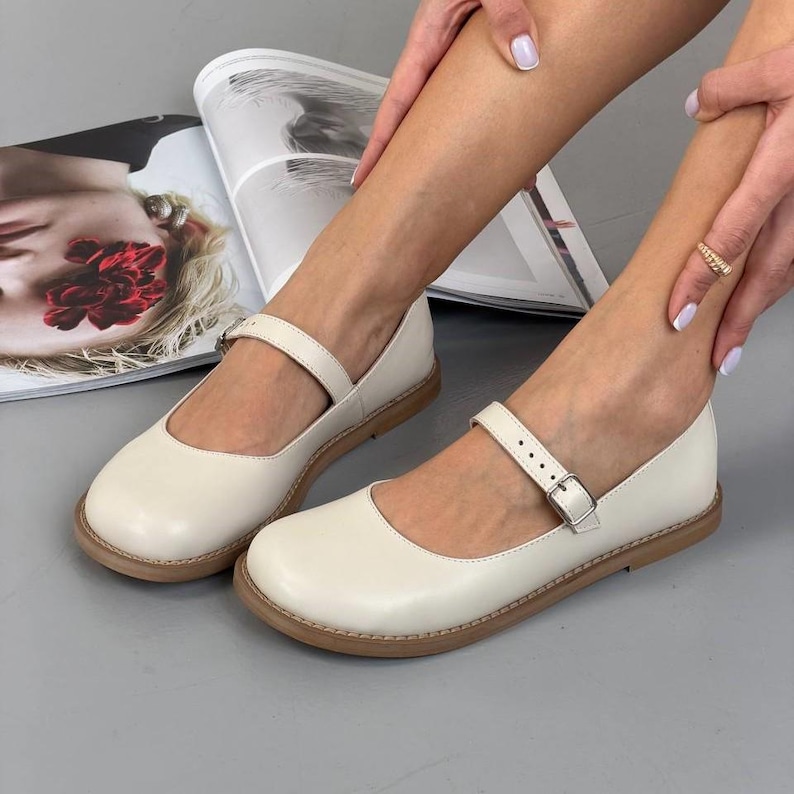 May include: Cream Mary Jane shoes with a rounded toe and a strap with a buckle. The shoes have a flat sole with a light brown trim. The shoes are displayed on a grey surface. The shoes are a classic style.