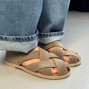 May include: A pair of taupe-colored sandals with a woven design. The sandals have a flat sole and are worn with blue denim jeans. The sandals are a neutral color and have a casual style.