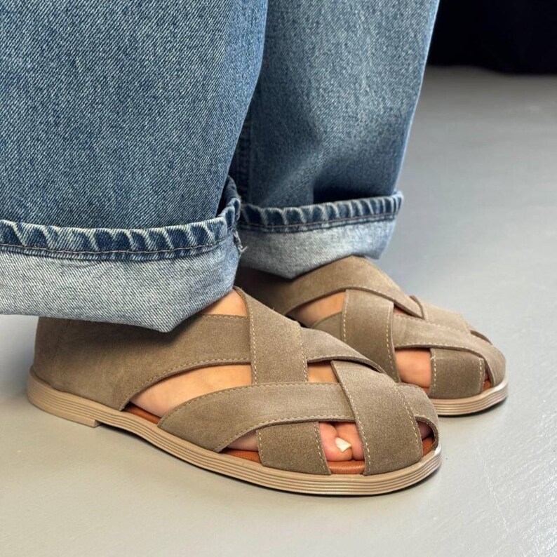 May include: A pair of taupe-colored sandals with a woven design. The sandals have a flat sole and are worn with blue denim jeans. The sandals are a neutral color and have a casual style.