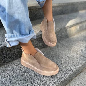 Puede incluir: Zapatos sin cordones de ante color canela con una suela gruesa de color marrón claro. Los zapatos tienen un diseño de corte bajo y se usan en escalones de piedra gris. Los zapatos son de color neutro.