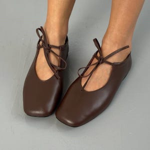 Puede incluir: Un par de bailarinas de cuero marrón con punteras cuadradas y lazos en los tobillos. Los zapatos tienen un diseño sencillo y elegante y están hechos de un material liso y mate. Las bailarinas se muestran sobre un fondo gris.
