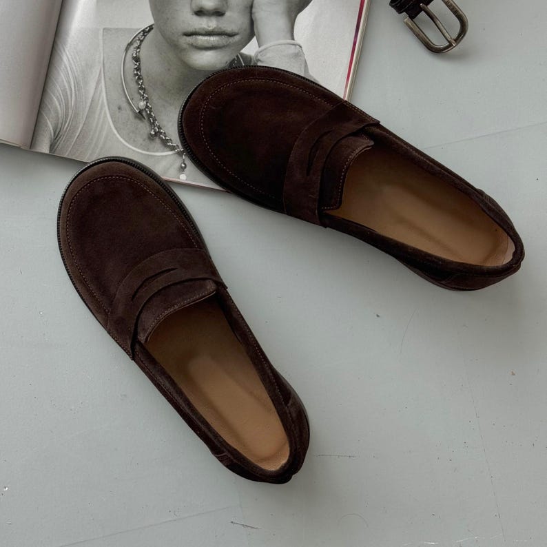 May include: A pair of dark brown suede loafers featuring a classic penny slot design across the vamp. The shoes have a rounded toe, a low profile, and a smooth interior lining. The exterior texture is matte and uniform in color throughout.
