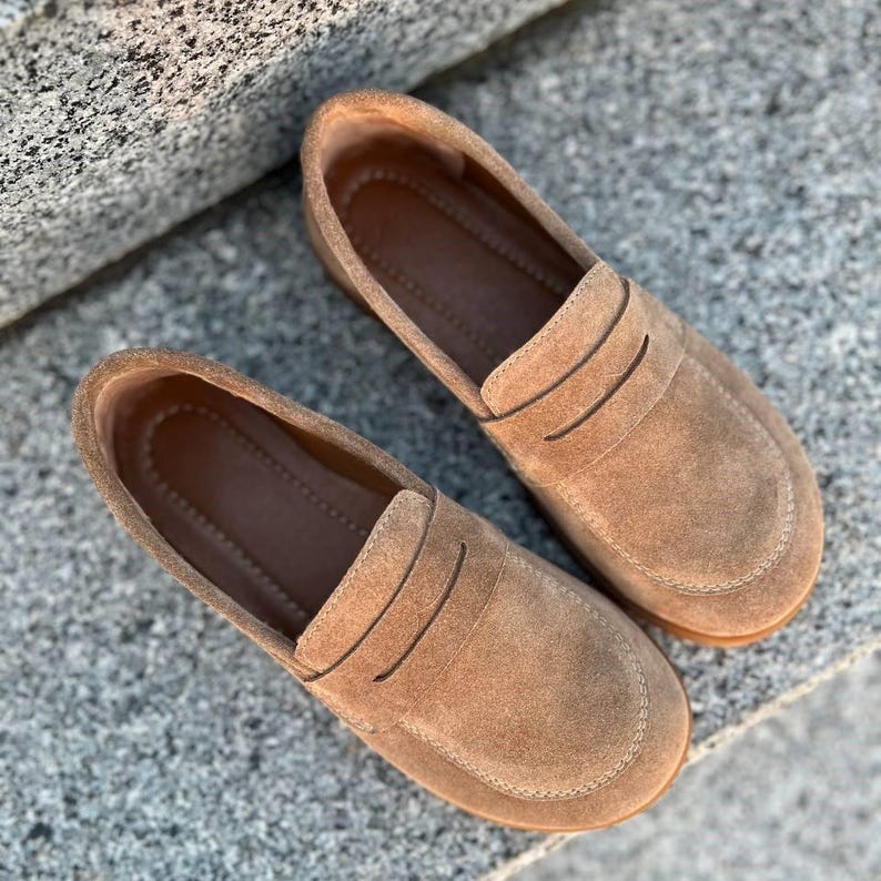 May include: A pair of tan suede loafers. The shoes have a rounded toe and a slip-on design. The upper features a decorative strap across the vamp. The soles are a darker brown. The shoes are placed on a textured gray surface.