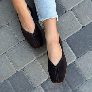 Pode incluir: Um par de sapatilhas de camur&ccedil;a castanho escuro com decote em V e bico quadrado. Os sapatos t&ecirc;m uma costura central. Usados com jeans azul claro, repousam sobre uma superf&iacute;cie de tijolos cinzentos. Cal&ccedil;ado elegante.